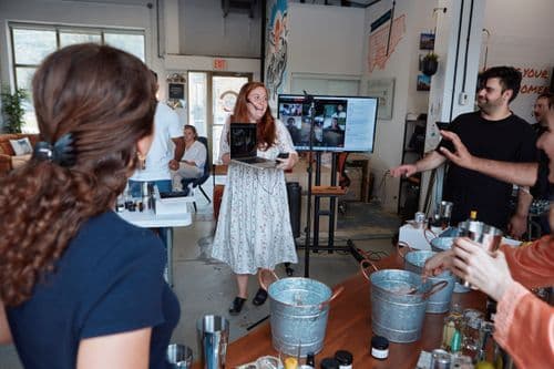 People in a workshop setting making cocktails, with a woman holding a laptop displaying a virtual meeting on a screen in the background.